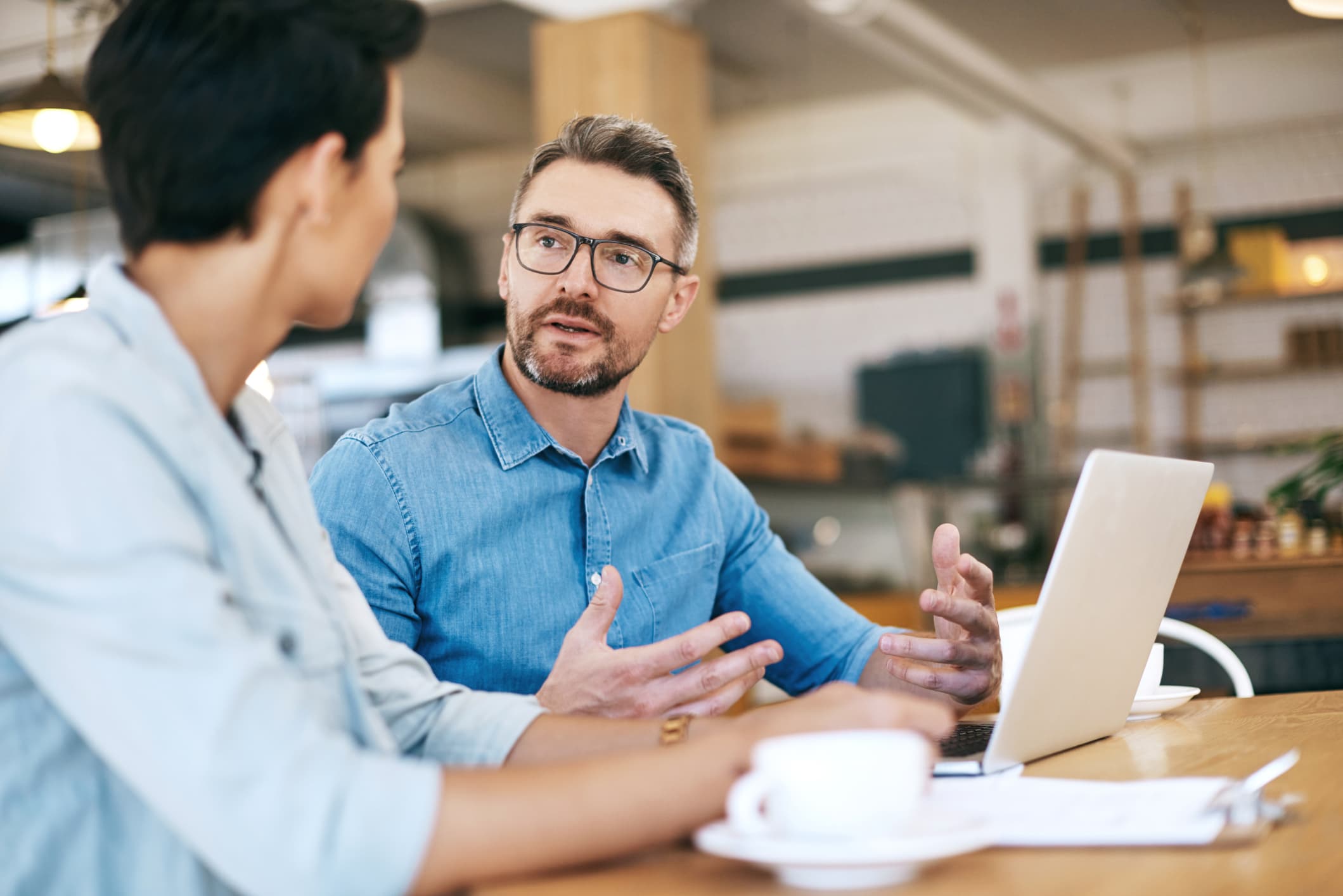 Man and woman sitting at a table, talking with each other with an open laptop.