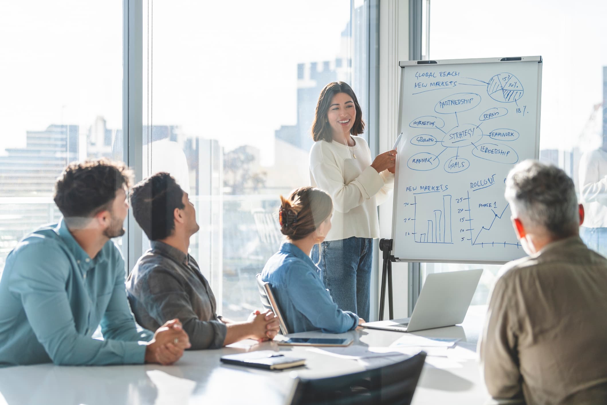 Four business people sitting at a table watching a woman present a strategy on a whiteboard.